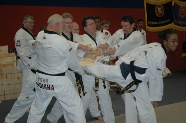 group of students in white gi holding board for woman in white gi kicking